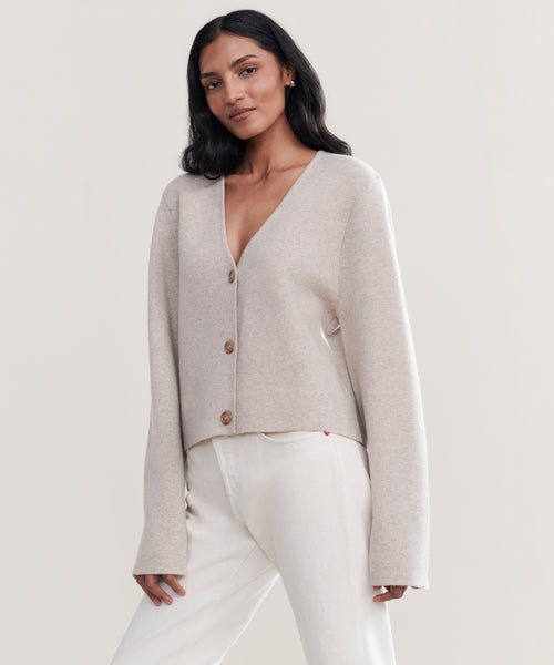A woman with long dark hair wears the Chelsea Cardigan in light beige with brown buttons and off-white pants, standing against a plain, light background.