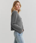 A woman with long, wavy blonde hair models the Chelsea Cardigan in gray with light blue jeans, standing sideways and looking at the camera against a plain light background.