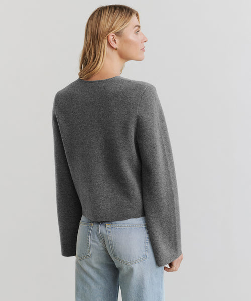 A person with light brown hair, wearing the Chelsea Cardigan in grey and light blue jeans, stands facing away from the camera against a plain light background.