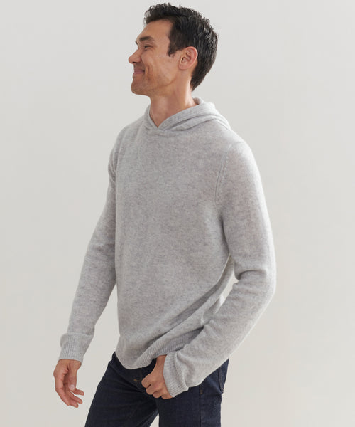 A man smiles while standing sideways in a light gray Mens Hoodie and dark jeans against a plain light background.