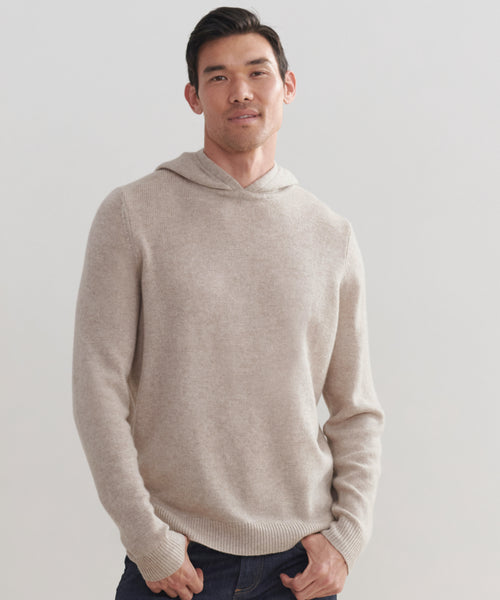 A man in a Mens Hoodie, light beige wool-cashmere, paired with dark jeans stands against a plain light background, smiling softly with his hands in his pockets.