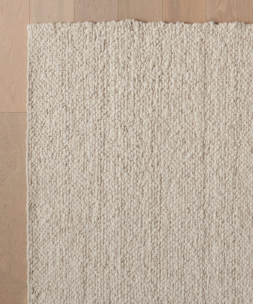 The Canyon Rug, a handmade, off-white wool piece from India with a chunky looped texture, is shown from above on a light wooden floor. The image captures a corner of this artisanal rug.