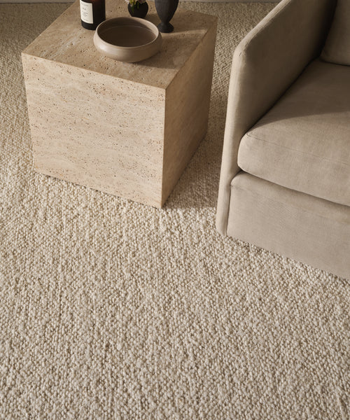 The Canyon Rug, a beige wool piece handmade in India, is placed beneath a stone square coffee table with decor, beside a light-upholstered armchair.