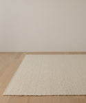 The Canyon Rug, a large, textured off-white wool piece handmade in India, rests on a light wood floor in a minimalist room with plain light-colored walls.