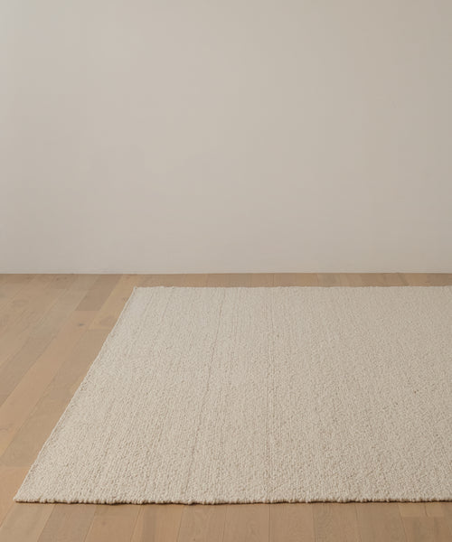 The Canyon Rug, a large, textured off-white wool piece handmade in India, rests on a light wood floor in a minimalist room with plain light-colored walls.