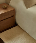 A wooden nightstand with a small bowl stands beside a neatly made bed with beige bedding and a matching pillow, next to the cream-colored Dune Rug from Lulu and Georgia on a dark wood floor.