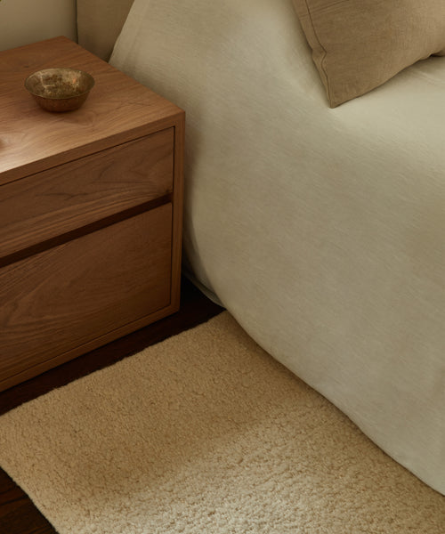 A wooden nightstand with a small bowl stands beside a neatly made bed with beige bedding and a matching pillow, next to the cream-colored Dune Rug from Lulu and Georgia on a dark wood floor.