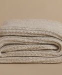 A folded knit blanket in a warm oatmeal beige color, showcasing its chunky ribbed texture against a solid beige background.