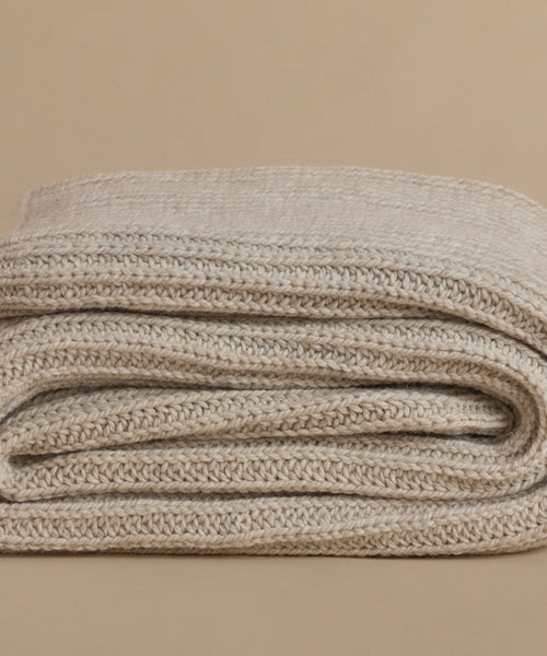 A folded knit blanket in a warm oatmeal beige color, showcasing its chunky ribbed texture against a solid beige background.