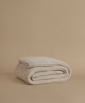 A folded knit blanket in a light oatmeal beige color, displayed on a solid, warm beige background.