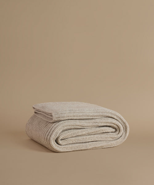 A folded knit blanket in a light oatmeal beige color, displayed on a solid, warm beige background.