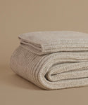 A folded chunky knit throw blanket in a warm oatmeal beige color, displayed on a matching solid beige background.