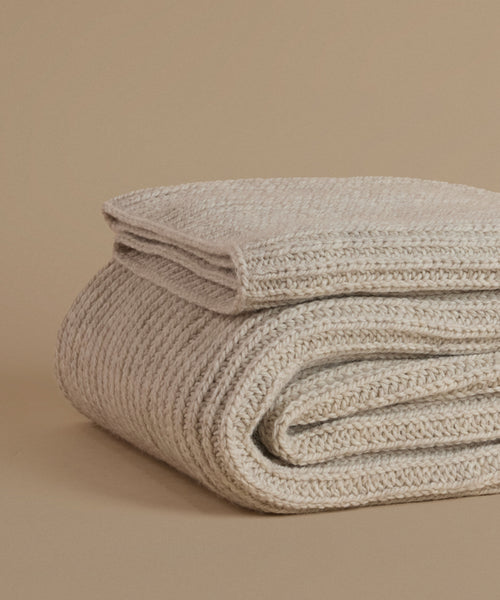 A folded chunky knit throw blanket in a warm oatmeal beige color, displayed on a matching solid beige background.