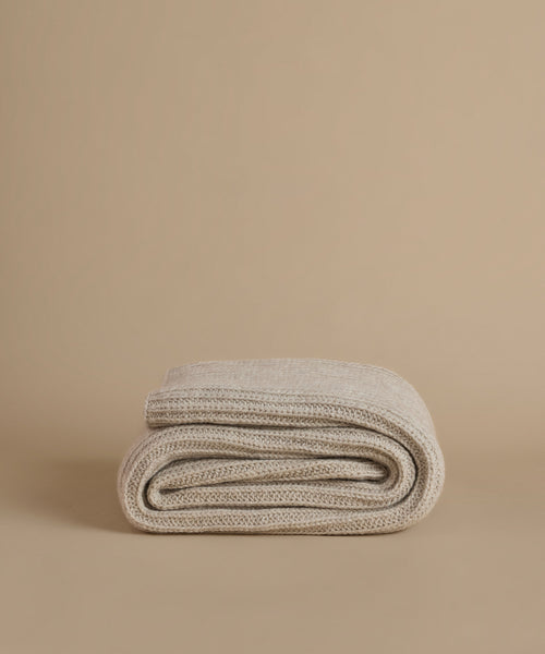 A folded knit blanket in a light oatmeal beige color, displayed on a matching solid beige background.
