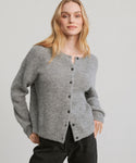 A woman with long blonde hair wears the Hayes Crewneck Cardigan in grey with black pants, standing against a plain light background and looking confidently at the camera.