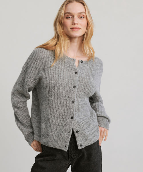 A woman with long blonde hair wears the Hayes Crewneck Cardigan in grey with black pants, standing against a plain light background and looking confidently at the camera.