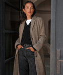 A woman stands confidently in a doorway wearing the Hyde Coat over a black sweater and white shirt, paired with black jeans, a belt, and her hands in her pockets.