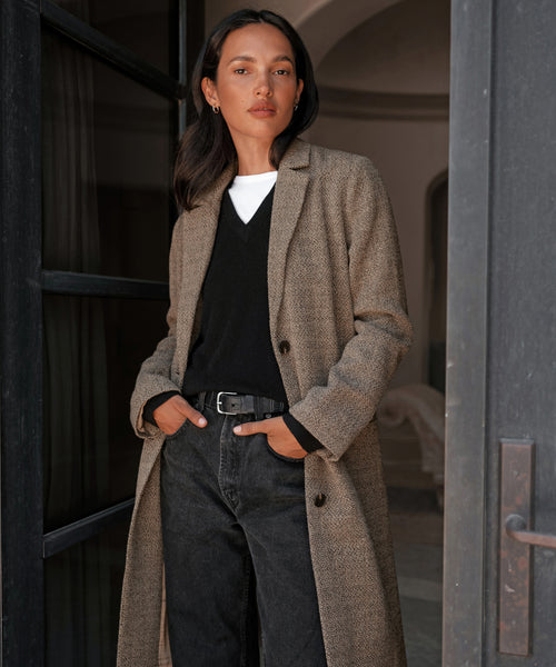 A woman stands confidently in a doorway wearing the Hyde Coat over a black sweater and white shirt, paired with black jeans, a belt, and her hands in her pockets.