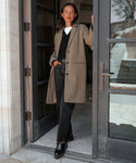 A woman stands in a doorway wearing the Hyde Coat over a black sweater and white top, paired with black jeans and boots. She has straight dark hair and leans against the door frame, looking ahead.