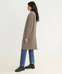 A woman with long dark hair stands in profile against a plain light background, wearing the knee-length tailored Hyde Coat, blue jeans, and black boots.