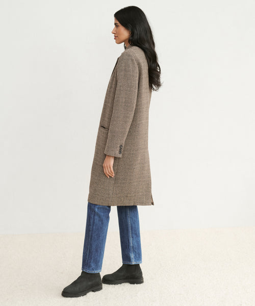 A woman with long dark hair stands in profile against a plain light background, wearing the knee-length tailored Hyde Coat, blue jeans, and black boots.