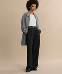 A woman with short curly hair and a confident expression stands against a beige background wearing the Wool Jones Trouser, paired with a gray coat and white top. Her hands are in her pockets, showcasing this modern, versatile pant inspired by Italian suiting.