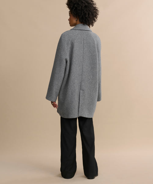 A person with short curly hair stands against a beige background, wearing a loose-fitting gray coat and Wool Jones Trouser, facing away from the camera.