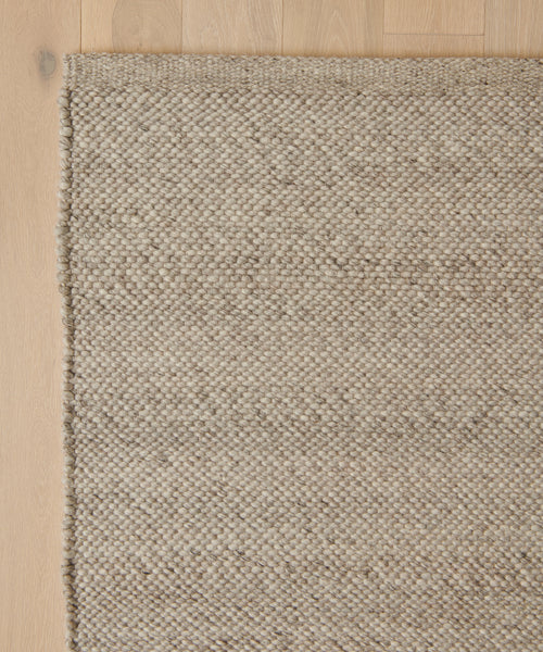 A close-up of the Lodge Handwoven Rug’s corner shows its textured beige New Zealand wool and subtle pattern, with a slightly raised edge on a light wood floor.