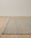 The Lodge Handwoven Rug in beige covers part of the corner space on a light wooden floor, set against an off-white wall in this minimalist room.