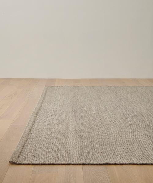The Lodge Handwoven Rug in beige covers part of the corner space on a light wooden floor, set against an off-white wall in this minimalist room.
