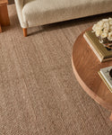 A close-up of a textured Rug Swatch in beige, paired with a round walnut coffee table displaying books and white roses, beside a light-colored upholstered couch.