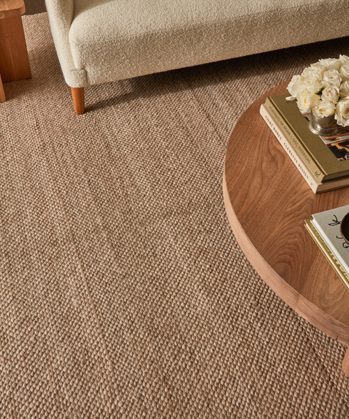 A close-up of a textured Rug Swatch in beige, paired with a round walnut coffee table displaying books and white roses, beside a light-colored upholstered couch.