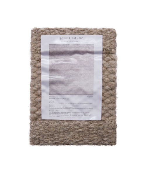 A Rug Swatch of chunky, handwoven beige wool with a Jenni Kayne product info sheet attached to the front, displayed against a plain white background.