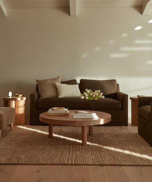 A cozy living room featuring a dark brown sofa, matching armchair, round wooden coffee table with books and white tulips, the Malawi Rug—hand-knotted wool and handmade in India—and soft sunlight streaming onto neutral walls.