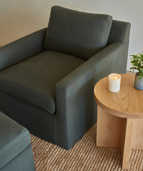 A dark gray armchair sits on the Malawi Rug, a sustainable, hand-knotted wool rug handmade in India, beside a light wooden round table with a lit white candle and small vase of green flowers.