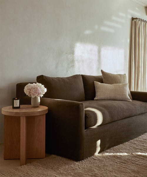 A dark brown sofa with beige pillows sits against a light wall. The handmade Malawi Rug from India grounds the space, while sunlight and shadows add warmth. A wooden side table displays a vase of pale flowers and a candle.