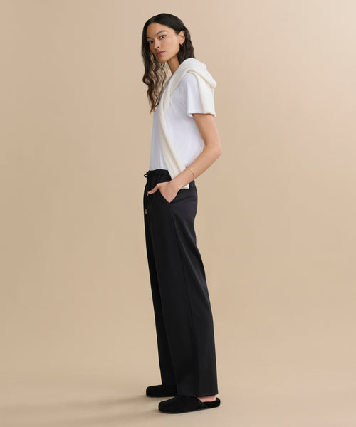 A woman stands in profile against a beige background, wearing a white t-shirt, the Wool Paloma Pant with an elastic drawstring waist, black slippers, and a white sweater draped over her shoulders. Her hands are in her pockets and her hair is loose.
