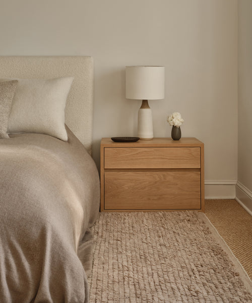 A cozy bedroom features a beige bed with neutral pillows, a wooden nightstand with a white lamp, flowers in a vase, and a decorative dish. Beside the bed, the Winward Rug adds warmth and texture to the floor.