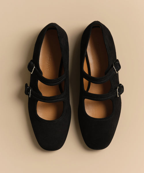 The Wren Flat, a pair of black suede Mary Jane shoes with double straps and rounded square toe, is photographed from above on a beige background.
