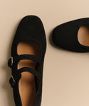 A close-up of two black suede Wren Flat Mary Jane shoes with double straps, rounded square toes, and silver buckles, shown on a beige background.