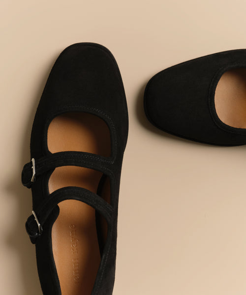 A close-up of two black suede Wren Flat Mary Jane shoes with double straps, rounded square toes, and silver buckles, shown on a beige background.
