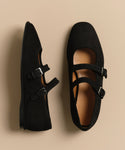 The Wren Flat, a pair of black suede Mary Jane shoes with double straps and rounded square toes, is displayed on a beige background—one shoe shown from the side, the other from above.
