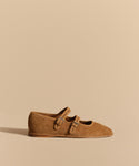 The Wren Flat, a tan suede Mary Jane with a rounded square toe and two thin buckle straps, is displayed on a neutral beige background with a soft shadow to its left.