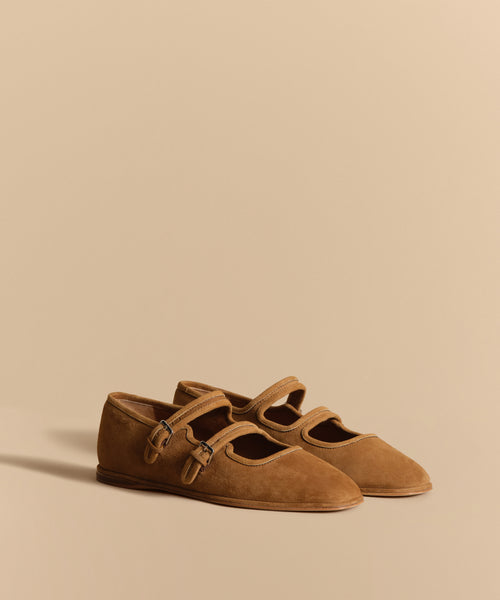 The Wren Flat is a tan suede Mary Jane with double straps, small buckles, and a rounded square toe, shown against a matching beige background.