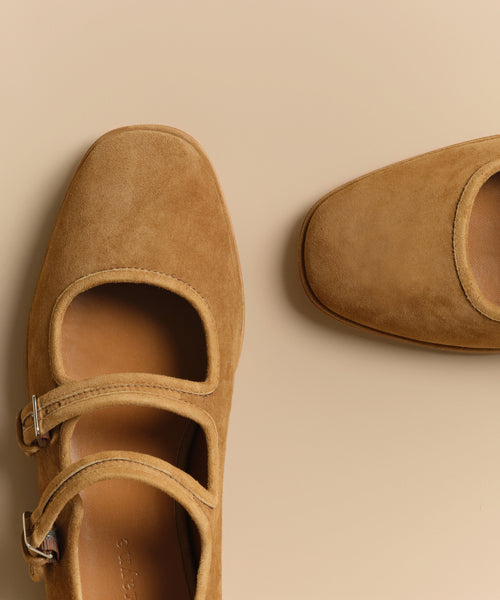 The Wren Flat in tan suede, featuring double straps and rounded square toes, is shown partially on a beige surface.