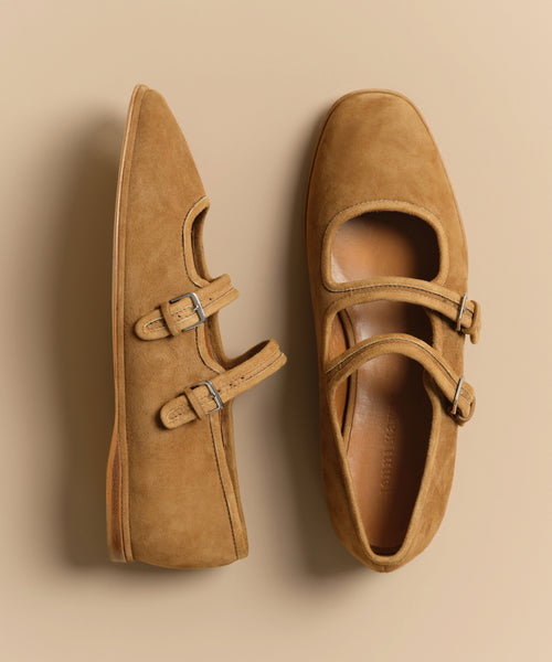 The Wren Flat, a pair of tan suede double-strap flats with buckle accents and rounded square toes, is shown on a beige background—one shoe displayed from the top, the other from the side.