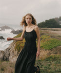 Woman in a black wynn dress standing on a coastal landscape with ocean and trees in the background.