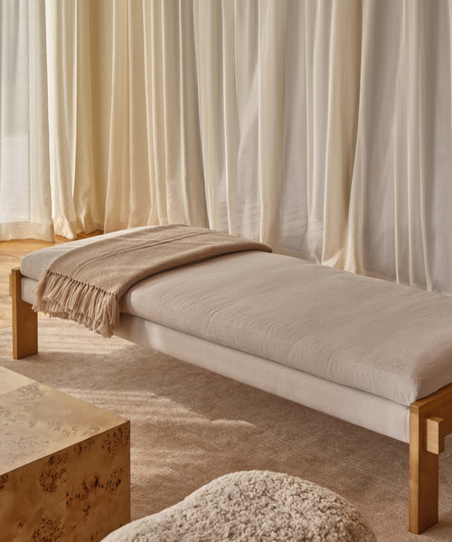A minimalist room features the Ynez Bench with a beige cushion and tan throw on a neutral rug by sheer cream curtains. A textured ottoman and light wood table complete the tranquil space.
