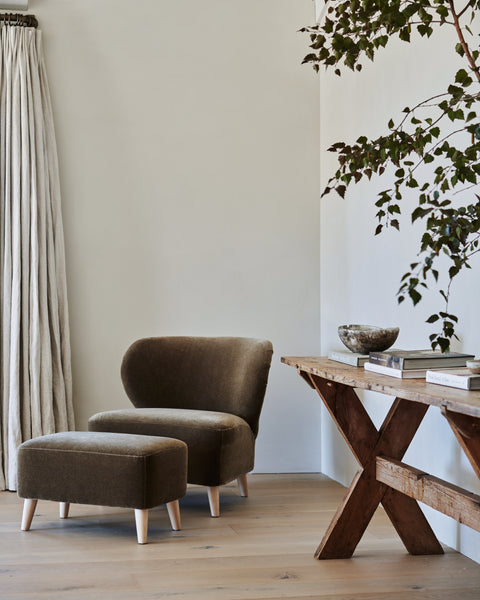 A cozy nook features a brown velvet armchair with the Brentwood Ottoman, rustic wooden table topped with books and a bowl, light wood floors, neutral walls, beige curtains, and leafy greenery—embodying inviting upholstered accent furniture style.