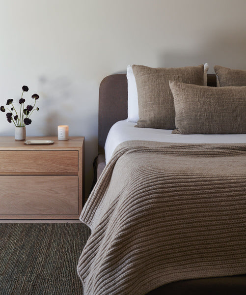 A modern bedroom showcases a neatly made Cove Bed in charcoal linen with textured brown pillows and a knitted beige blanket. A wooden nightstand holds a vase of dark flowers, candle, and tray, adding to the cozy, minimalist vibe.